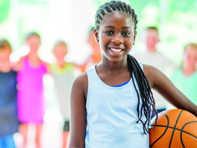 A multi-ethnic group of elementary age students are playing together in the gym during recess. One girl is holding a basketball and is smiling while looking at the camera.