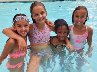 Image of young girls smiling and posing together in pool having fun at Coppermine 4 Seasons Healthclub in Hampstead Maryland. Master the water with Coppermine's swimming classes, catering to all skill levels and ages. Caption: Find the best indoor swimming pool near you at Coppermine, with heated pools and swimming programs.